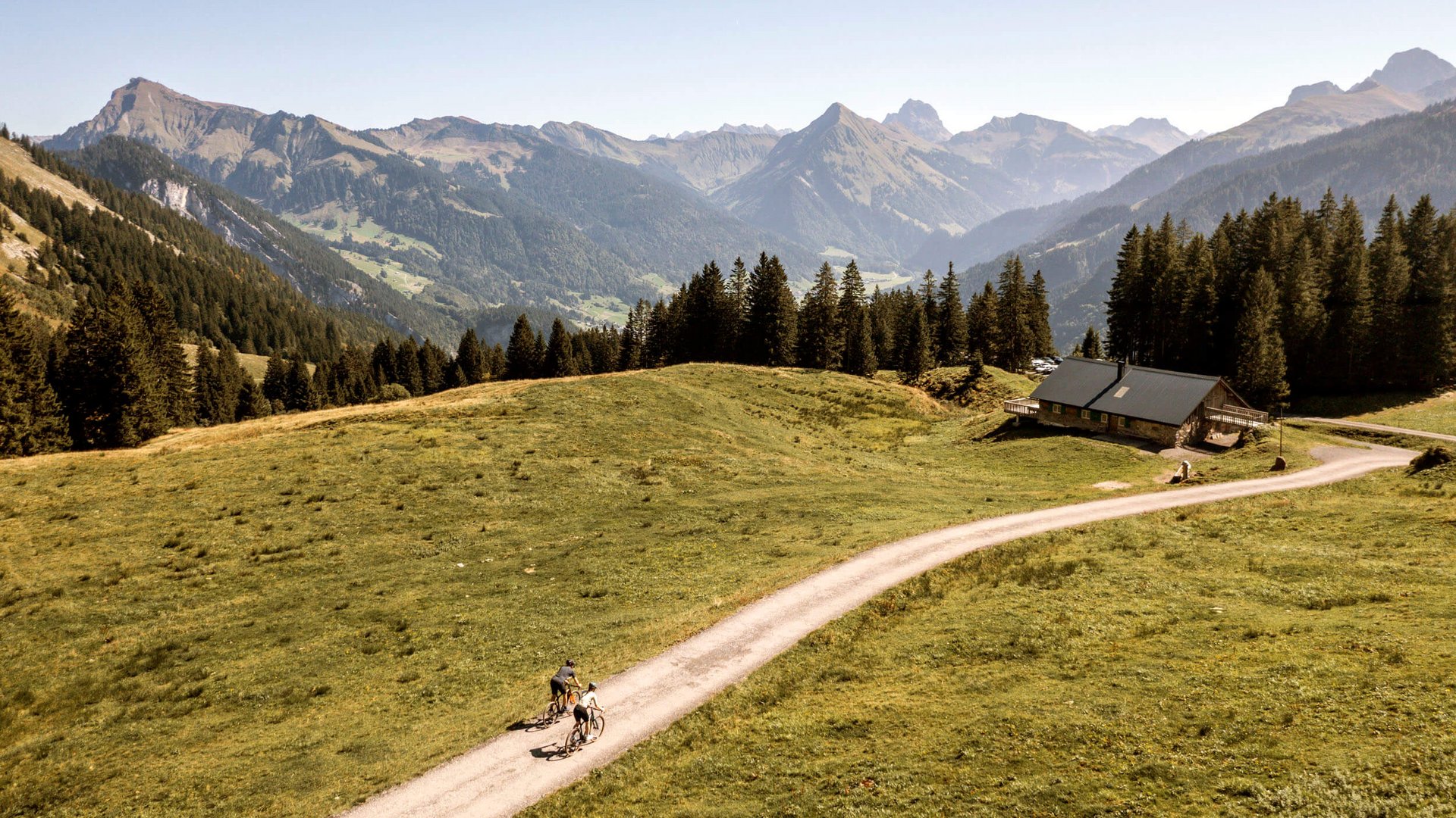 Zwei Radfahrer auf einem Weg über eine Almwiese mit Berghütte und Alpenpanorama (© Heiko Mandl)
